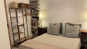A bedroom with a bed, a wooden wardrobe, a hanging shelf, a lamp, and a stone wall.