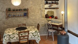 A rustic dining area with a stone wall, wooden beams, a tiled floor, a table with a patterned tablecloth, chairs, a hanging light fixture, and a stove.
