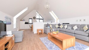 A modern loft apartment with a dining area, a living room with a grey sofa, and a kitchen in the background. - 3 Bed in Bideford (oc-75539) (Northam)
