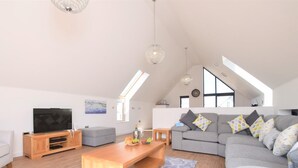 A spacious living room with a large grey sofa, wooden coffee table, and a flat-screen TV. - 3 Bed in Bideford (oc-75539) (Northam)