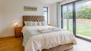 A bedroom with a large bed, a nightstand, a lamp, a painting on the wall, and a sliding glass door leading to a garden. - 3 Bed in Bideford (oc-75539) (Northam)