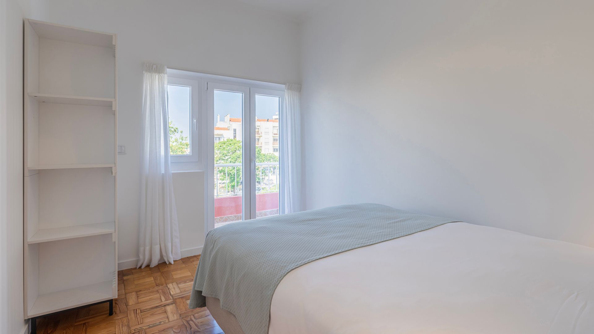 A bedroom with a bed, a wardrobe, a balcony with a view of buildings, and a window with curtains.