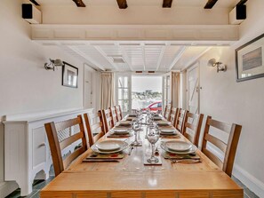 A long dining table set for a meal with chairs on both sides, a view of the sea through large windows, and a white ceiling with exposed beams. - 6 Bed in Fowey (oc-75581) (Fowey)