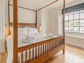 A four-poster bed with white linens, a wooden floor, and a window with a view of a town. - 6 Bed in Fowey (oc-75581) (Fowey)