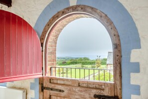 Blick durch ein rundbogiges Fenster mit rotem Rollo, der einen Balkon mit Metallgeländer und ein Gebäude im Hintergrund zeigt.