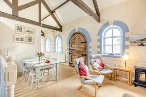 Ein Zimmer mit Holzbalkendecke, Bogenfenstern, einem Esstisch mit Stühlen und einem Kamin.