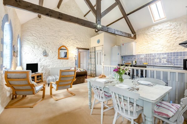 A room with a stone wall, wooden beams, a dining table with chairs, a sofa, a television, and a kitchen area with blue tiles.