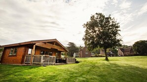 A wooden cabin with a porch, surrounded by a grassy lawn and a large tree. - 1 Bed in Worcester (oc-75600) (Halmond's Frome)