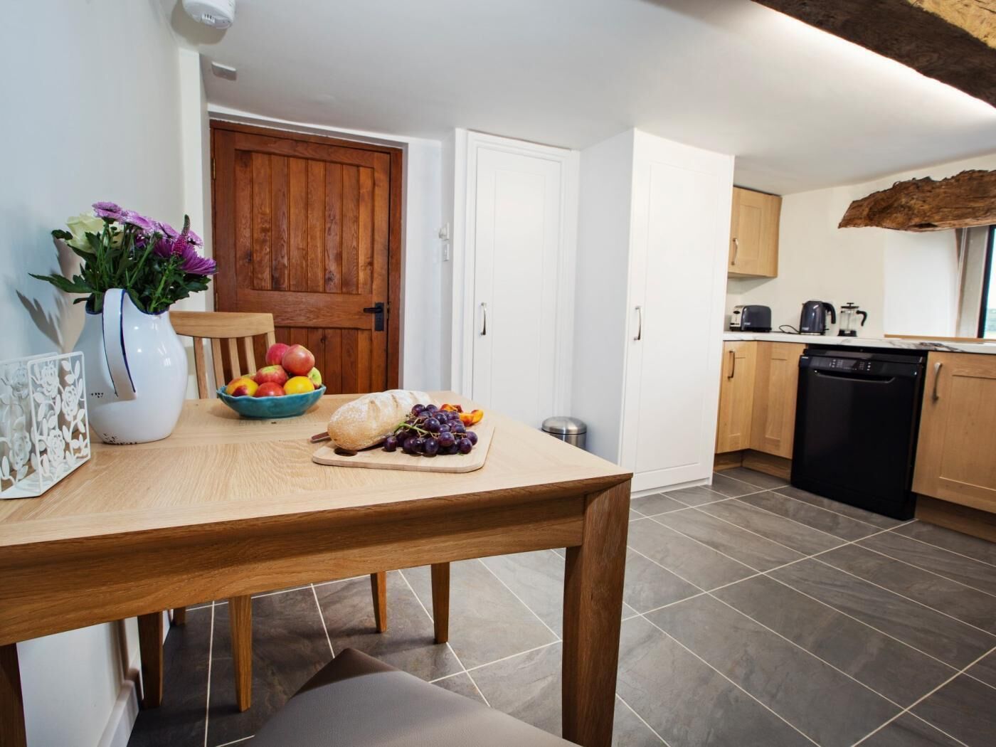 A kitchen with a wooden table, a bowl of fruit, a loaf of bread, and a vase with flowers.