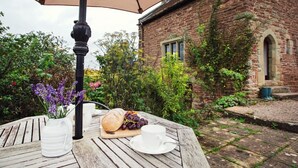 A round wooden table with a white pitcher, a loaf of bread, grapes, and a cup, under a beige umbrella, in front of a brick building with ivy. - 1 Bed in Worcester (oc-75600) (Halmond's Frome)