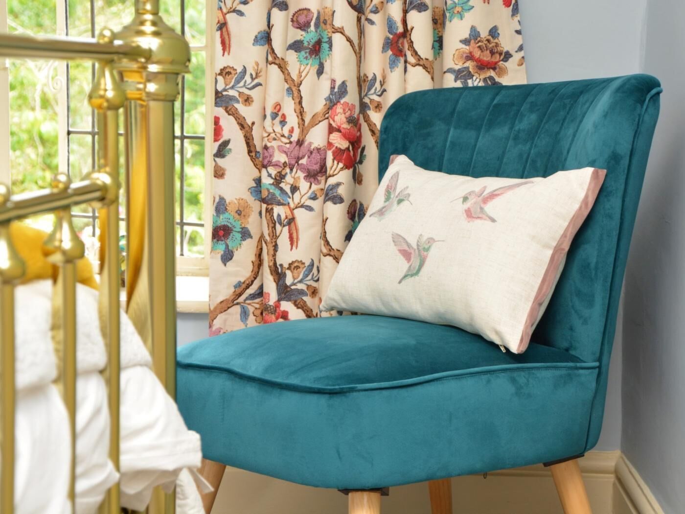 A teal armchair with a floral-patterned pillow, positioned near a window with a brass railing.