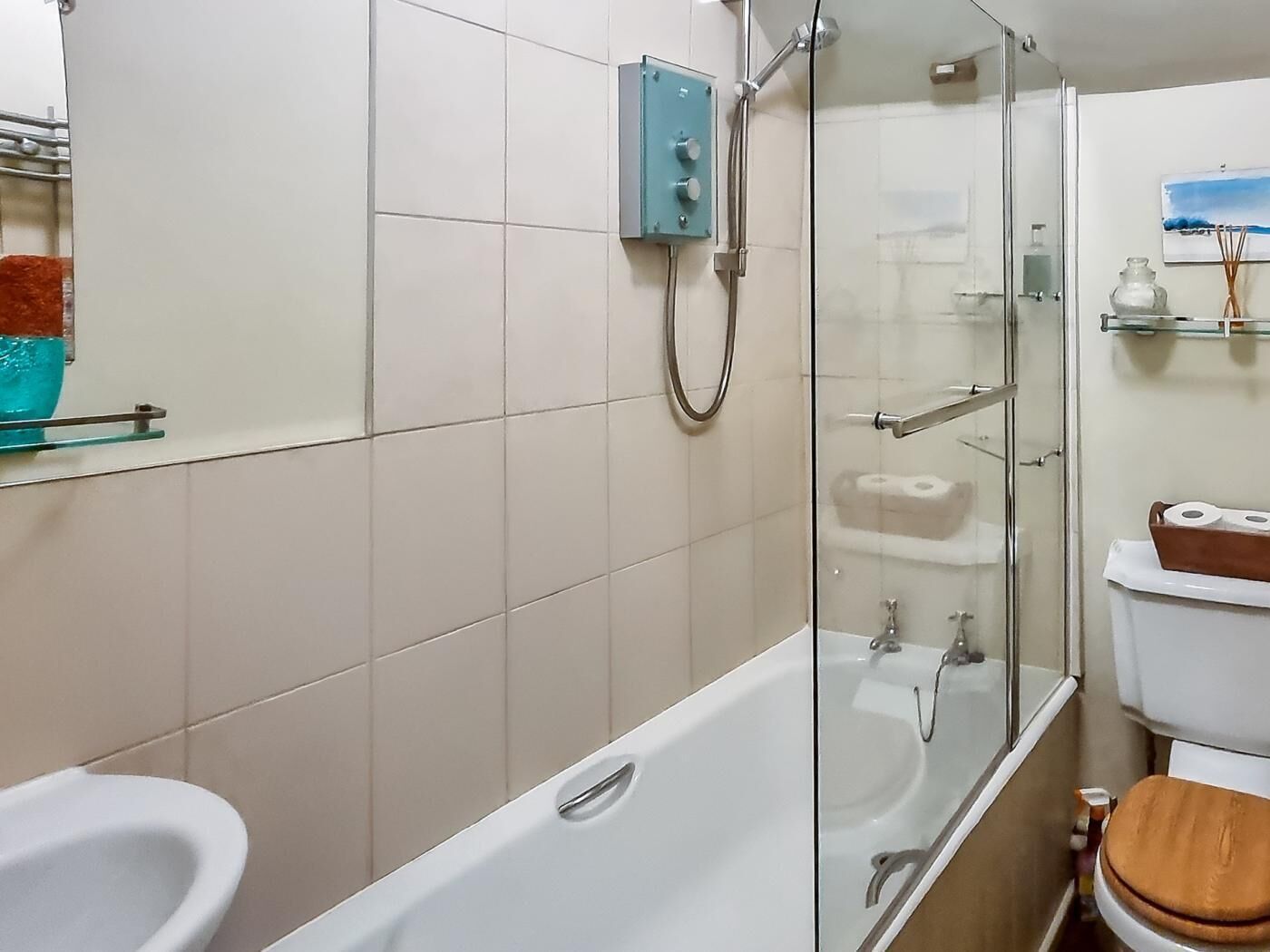 Une salle de bain avec une douche à parois de verre, une baignoire blanche, des toilettes avec un abattant en bois et une étagère en verre où sont rangés les articles de toilette.