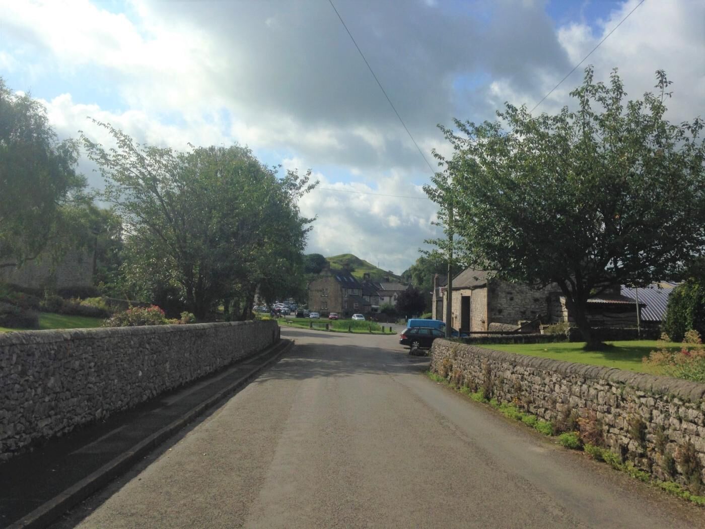 A rural road flanked by stone walls and trees, leading to a stone building with a thatched roof.