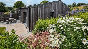 A rustic wooden structure with a patio area, surrounded by a variety of flowers and greenery. - 3 Bed in Penzance (oc-75635) (Gulval)