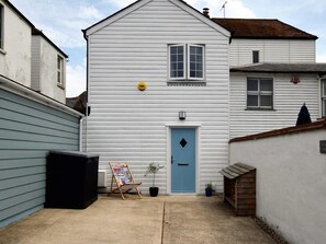 A white building with a blue door, a small garden with a chair and a plant, and a wooden structure to the right. - 3 Bed in Folkestone (oc-75607) (Sandgate)