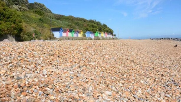 Uma praia de seixos com cabanas coloridas ao fundo.