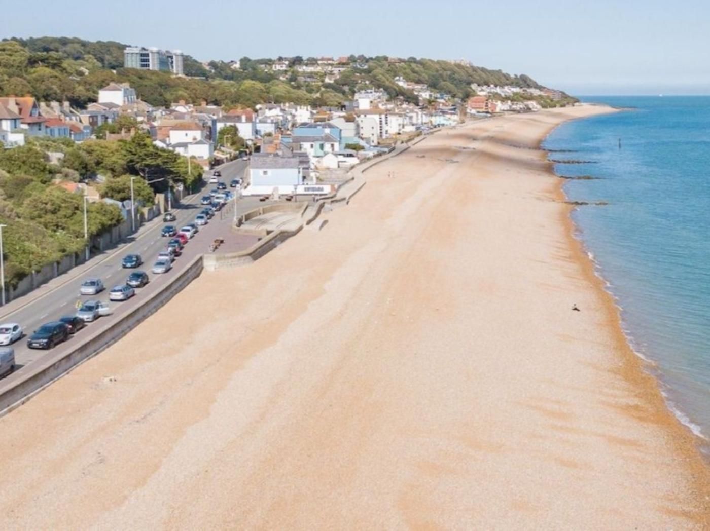A coastal road with parked cars, a beach, and buildings in the background.