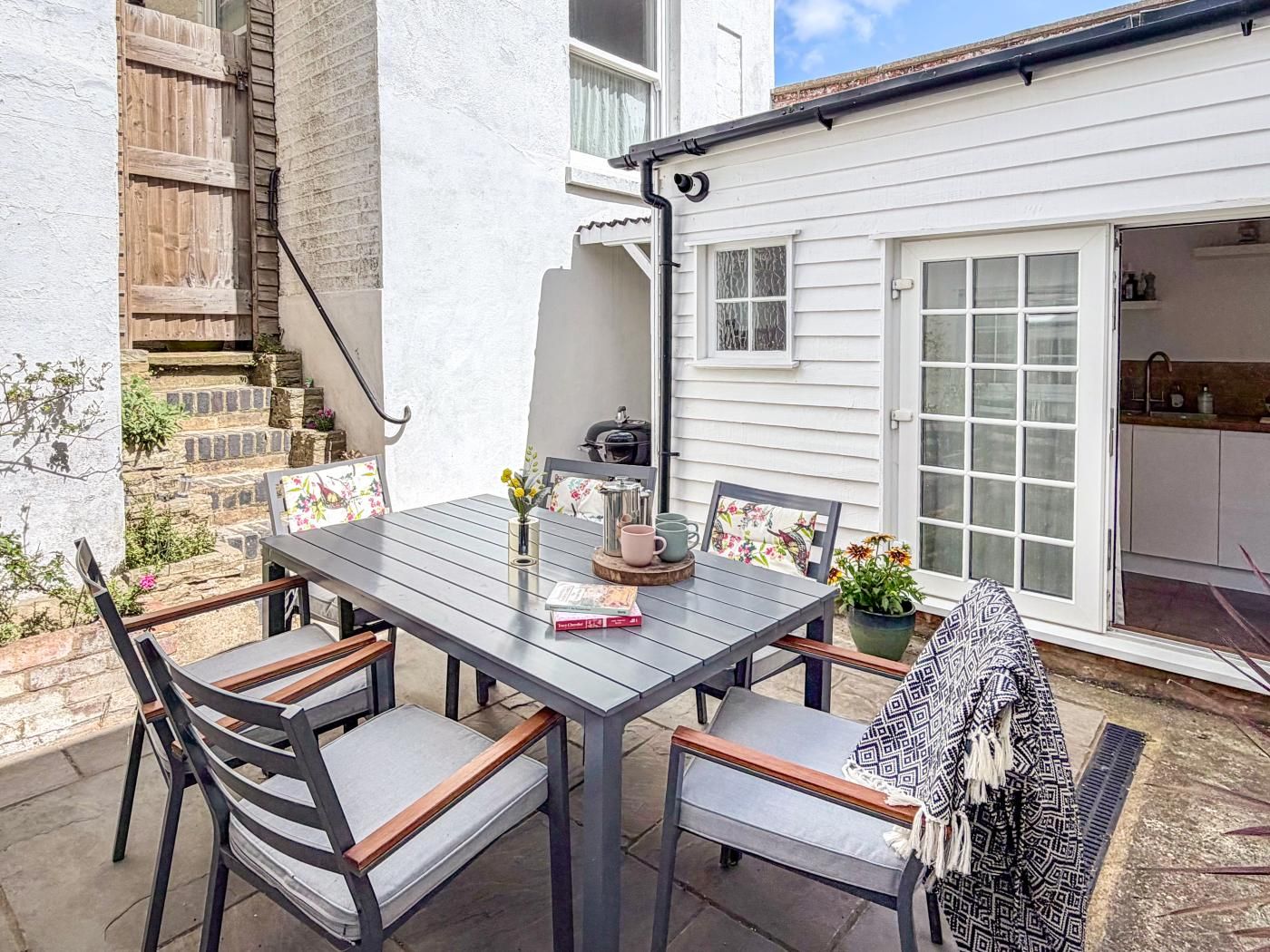 Eine Terrasse im Freien mit einem Holztisch und Stühlen, einem Blumentopf und Blick auf ein weißes Haus mit Garage.
