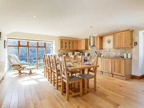 Eine geräumige Küche mit Holzausstattung, ein Essbereich mit Tisch und Stühlen sowie große Fenster mit Blick ins Freie.