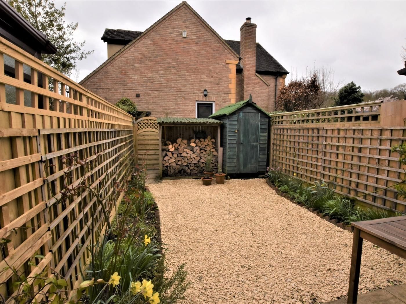A gravel pathway leading to a brick house with a green-roofed shed and a wooden fence.