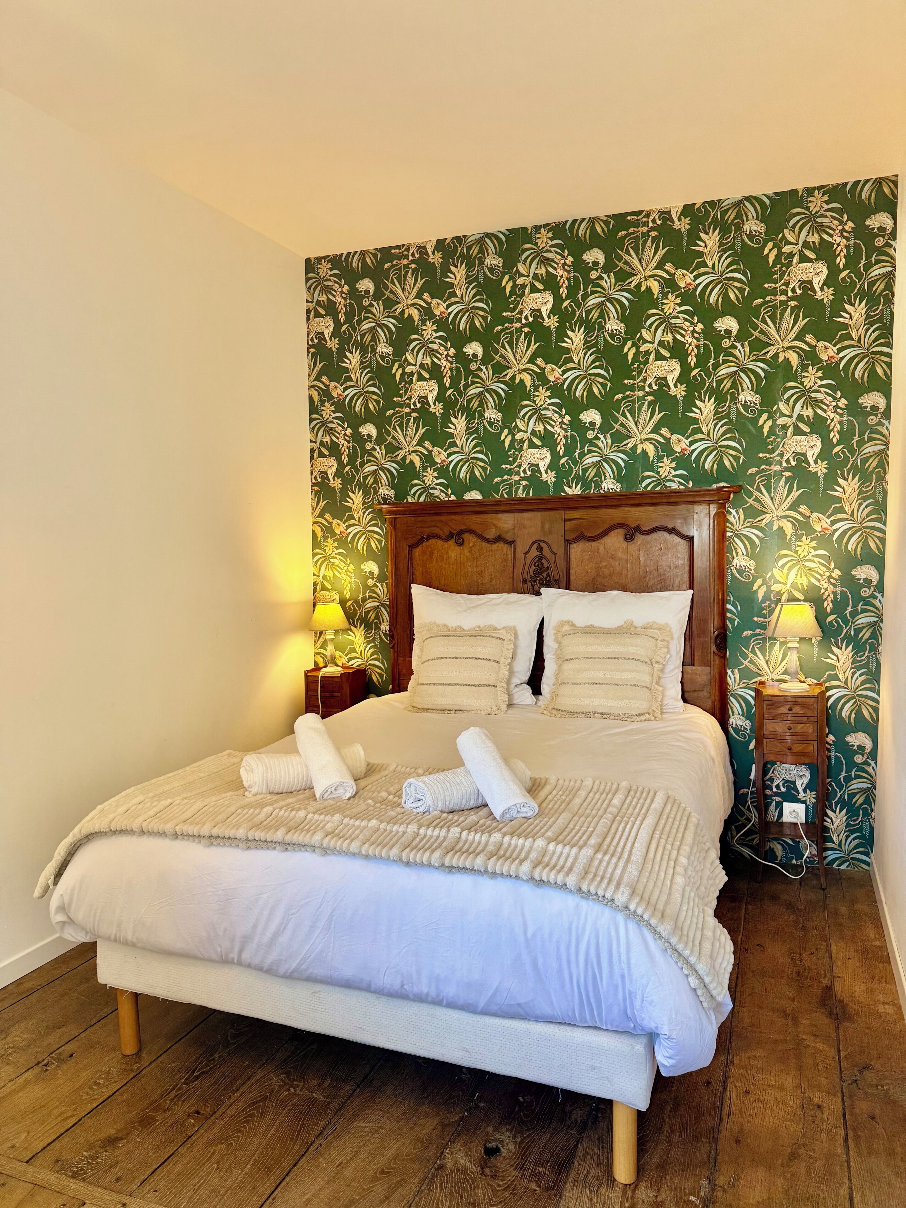 A bed with a wooden headboard, white bedding, and rolled towels. There are two bedside tables with lamps. The wall has a green floral wallpaper.