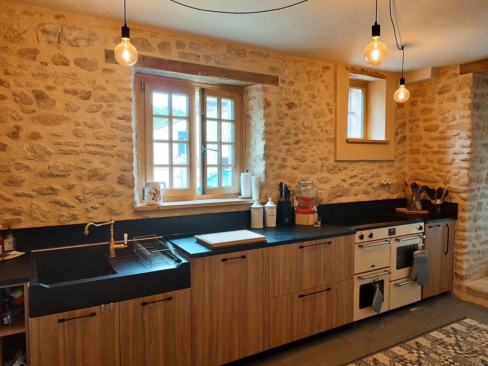 A kitchen with stone walls, wooden cabinets, a window, and hanging light bulbs.
