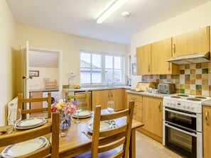 A compact kitchen with wooden cabinets, a dining table set for four, and a window providing natural light. - 3 Bed in Lairg (oc-ca026) (Laxford Bridge)