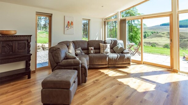 Un salon moderne avec un canapé d’angle, un plancher en bois et de grandes fenêtres donnant sur l’extérieur.