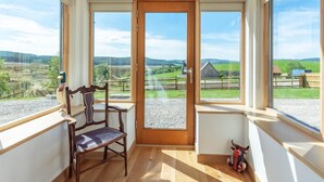 Une pièce avec un banc en bois, une porte donnant sur l’extérieur et de grandes fenêtres offrant une vue sur un paysage rural.