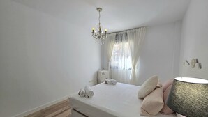 Un dormitori amb llit, un penjoll, una tauleta de nit, una làmpada i una finestra amb cortines.