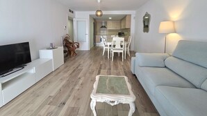 Un saló modern amb televisió de pantalla plana, un sofà, una taula de dinar amb cadires i una cuina al fons.