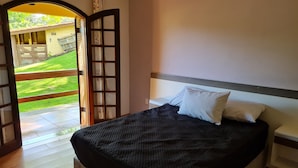 Un dormitorio con una cama, una mesita de noche y vistas a una zona verde a través de una puerta abierta.