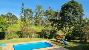Una piscina con borde de piedra, un tobogán y un columpio en un área verde con árboles.