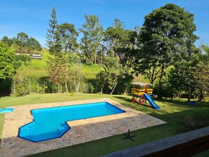 Piscina com borda de pedra, escorrega e baloiço numa área gramada com árvores.