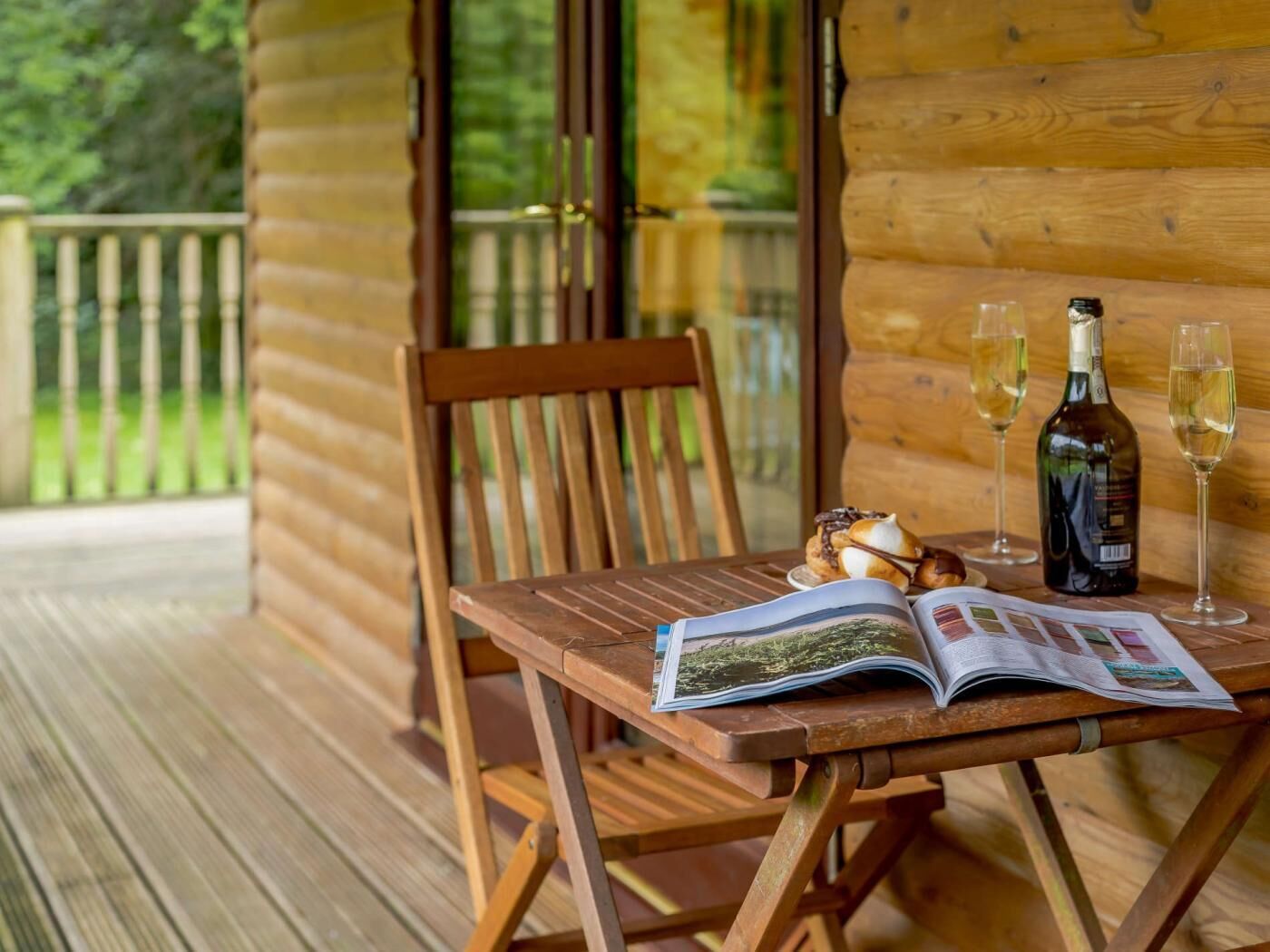 Une terrasse en bois avec une chaise en bois, une table sur laquelle se trouve un magazine, une bouteille de vin et deux verres.