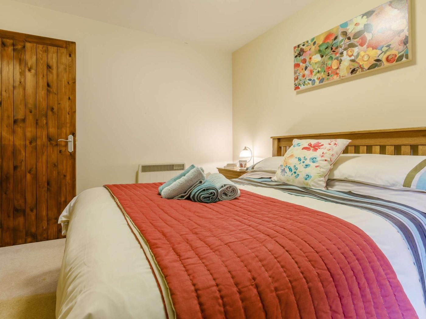 Un dormitorio con puerta de madera, cama con colcha roja y un cuadro floral en la pared.