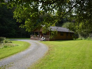 A wooden cabin with a porch, surrounded by a gravel path and grassy area. - 2 Bed in Holsworthy (oc-85562) (Bradworthy)