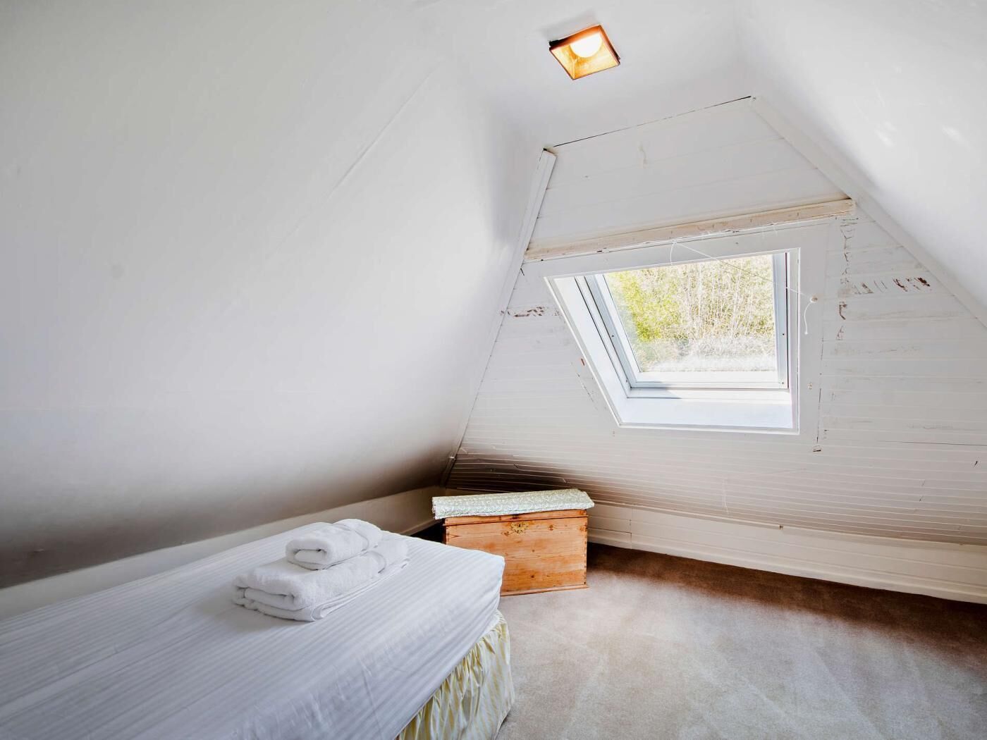 A small, white, attic-style bedroom with a skylight, a single bed with white linens, and a wooden chest.