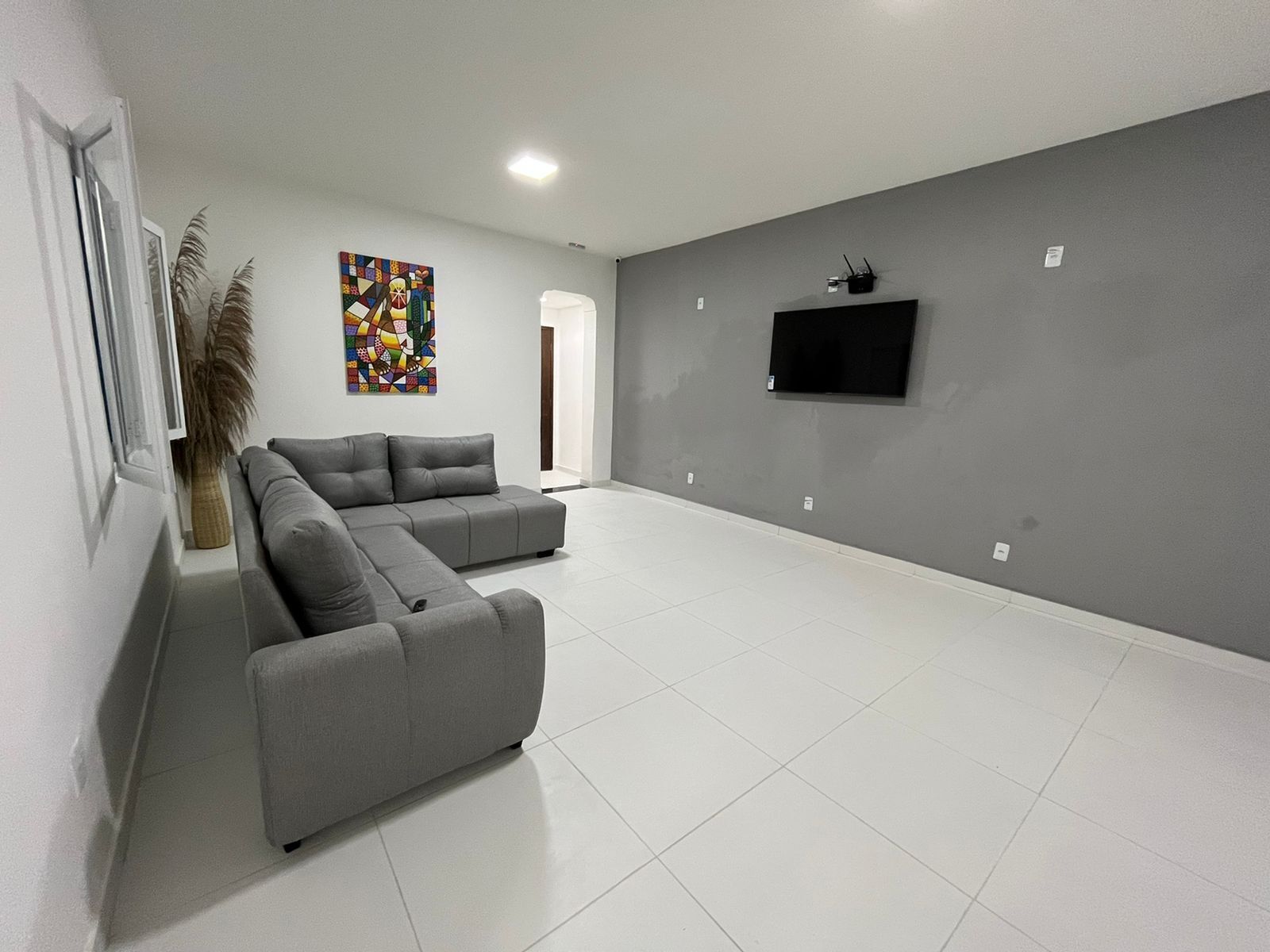 A modern living room with a grey sectional sofa, a flat-screen TV mounted on the wall, and a large window with a view of the outside.