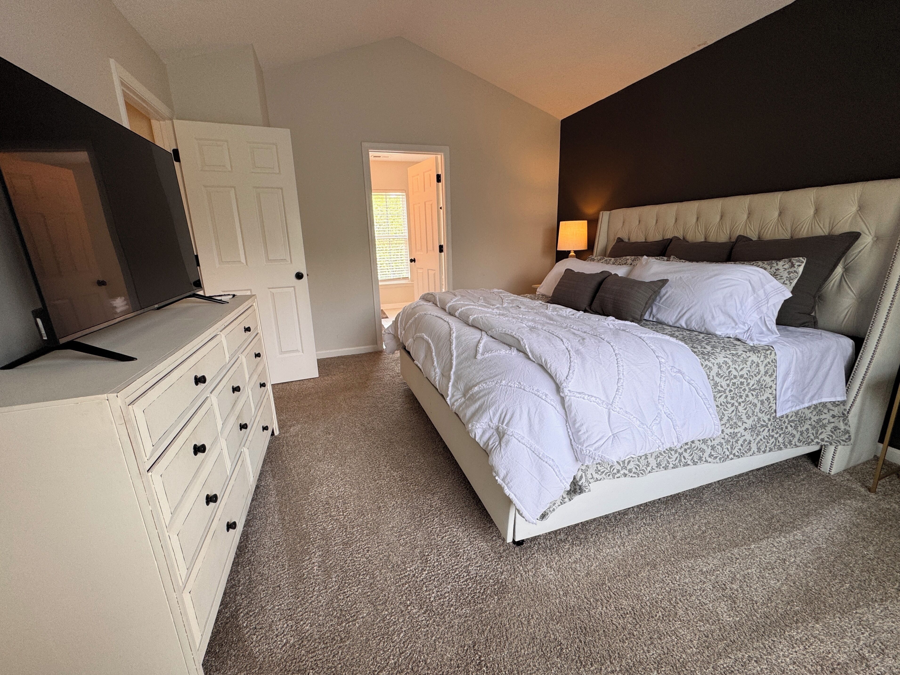 Une chambre à coucher comprenant un lit, une commode, un téléviseur et une porte donnant sur une autre pièce.