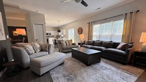 A spacious living room with a large sectional sofa, a coffee table, and a dining area with a kitchen in the background.