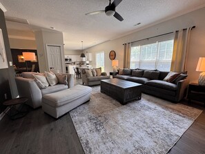 A spacious living room with a large sectional sofa, a coffee table, and a dining area with a kitchen in the background. - HOT TUB, Community Pool, AC, 5-Bedroom Oasis in Simpsonville (Simpsonville)