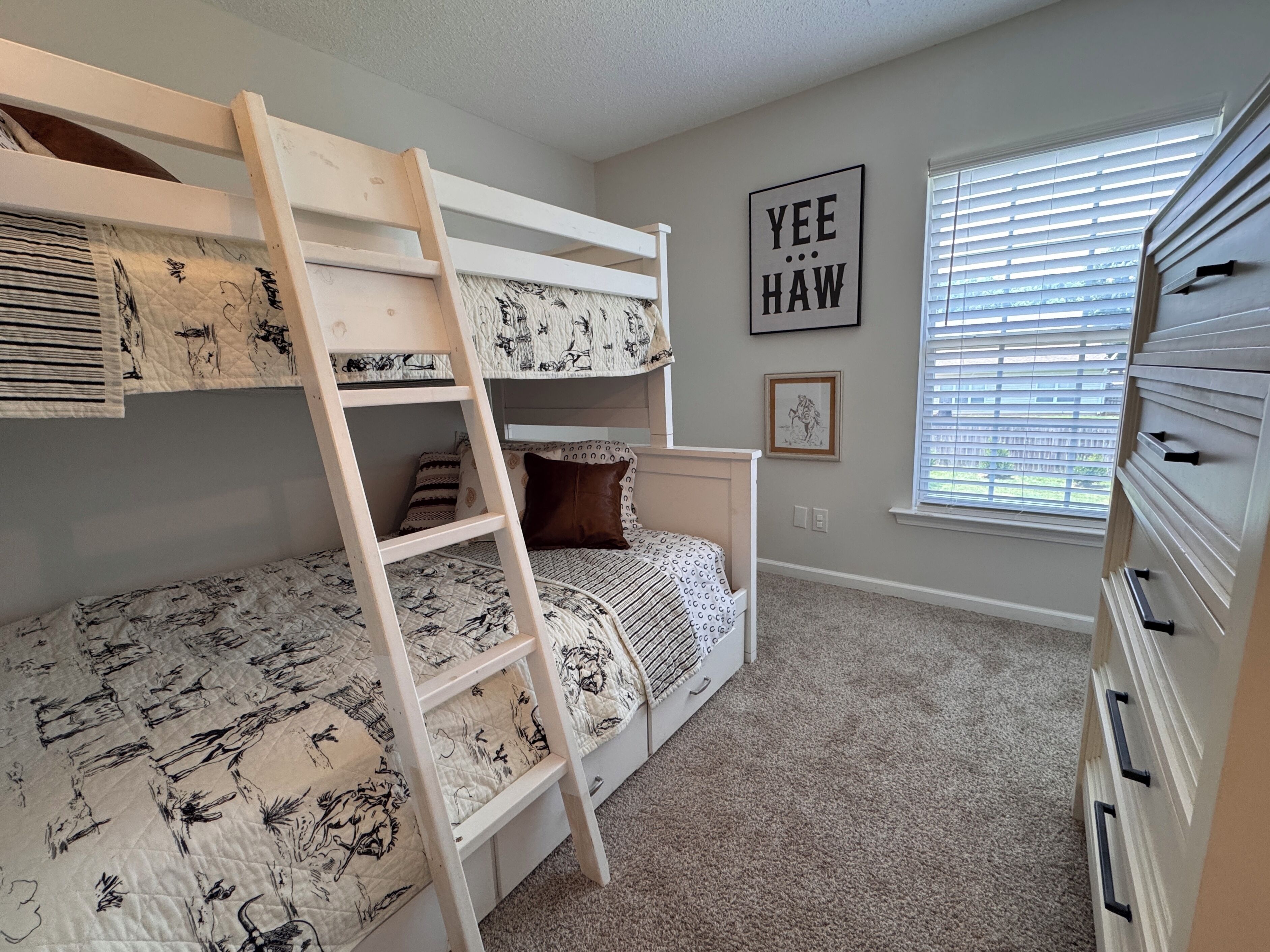 A bunk bed room with a ladder, a wall with a framed picture, and a window with blinds.