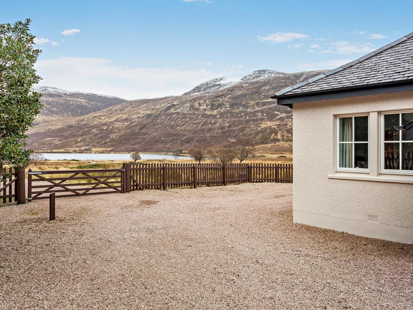 A building with a gravel driveway, a wooden fence, and a view of a lake and mountains.