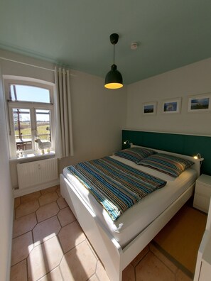 Une chambre à coucher avec un lit, une fenêtre, un radiateur, un luminaire au plafond et des tableaux encadrés accrochés au mur.