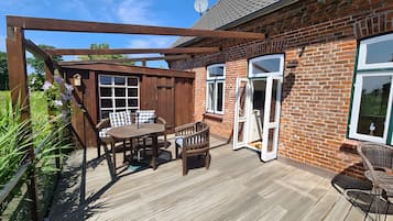 Eine Holzterrasse mit Tisch und Stühlen, einer Pergola und einem Ziegelhaus mit großen Fenstern und Türen.