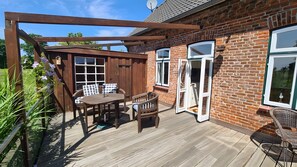 Une terrasse en bois avec une table et des chaises, une pergola et une maison en briques dotée de grandes fenêtres et de grandes portes.