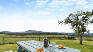 Ein Picknicktisch mit Kaffeekanne, Tasse und Croissant in einer grünen Wiese mit Baum und sanften Hügeln im Hintergrund.