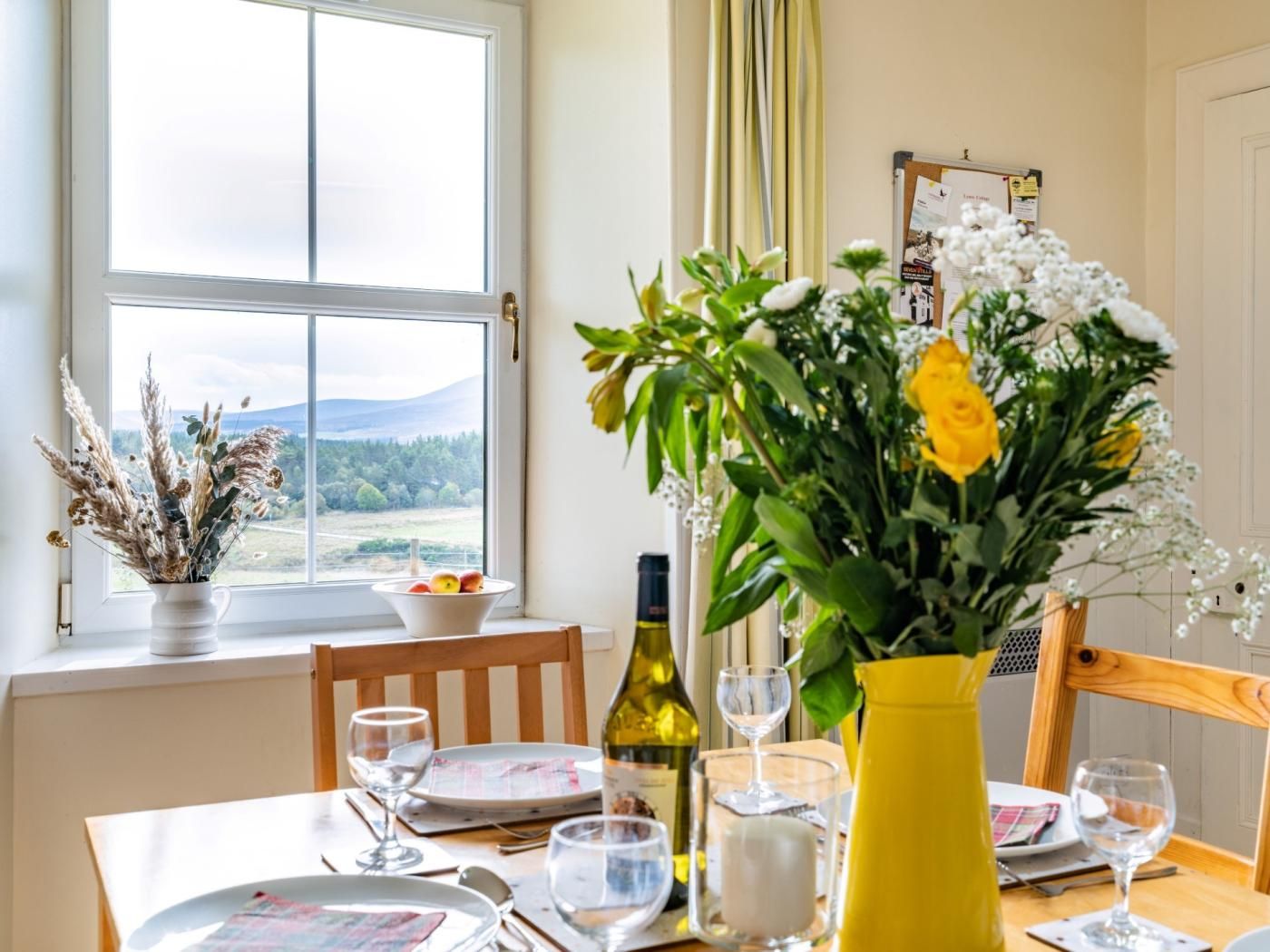 Une table à manger dressée avec des assiettes, des verres et une bouteille de vin, avec une fenêtre donnant sur un paysage.