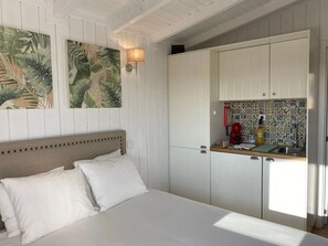 Un dormitorio con cama, almohadas blancas, cabecera con detalles circulares, una lámpara de pared y una zona de cocina con salpicadero de azulejos y un electrodoméstico rojo.